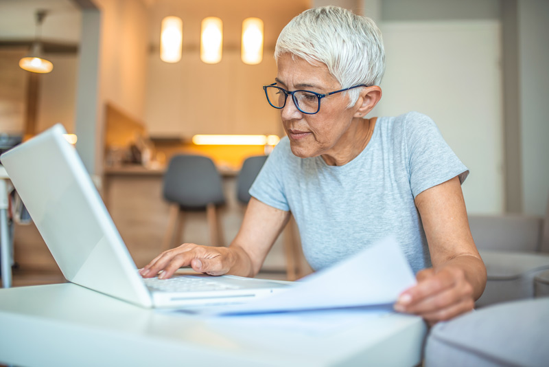 A short-haired senior woman, wearing glasses and looking curious as she gazes at her laptop.