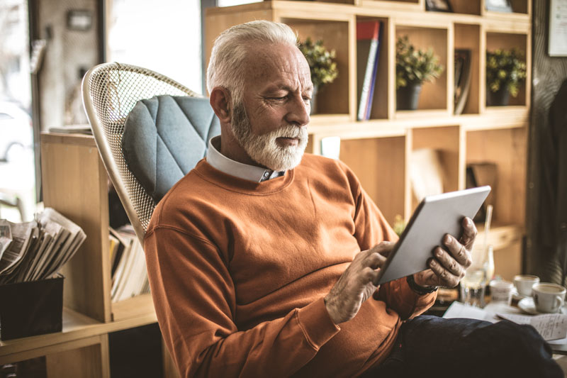 A senior man, wearing glasses, reads an article on his iPad.