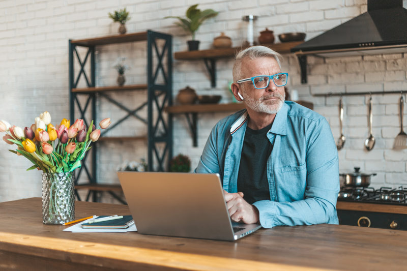 A senior man with glasses sitting in the kitchen with an open laptop, appearing curious as he looks away.