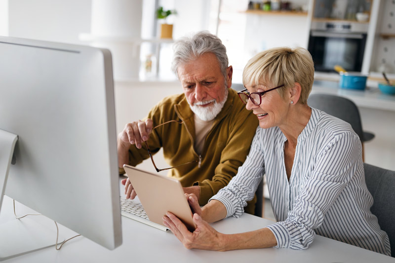 A senior couple, both wearing glasses, sits by a computer, discussing while referring to their notes.